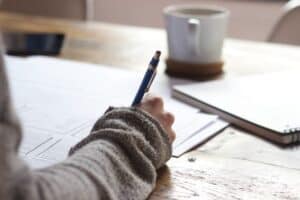 A person sits at a wooden desk, writing on a sheet of paper with a blue pen. Their sleeve indicates they're wearing a cozy, grey, knit sweater. In the background, a blurred mug of coffee sits next to a notebook, suggesting a focused work environment. The setting exudes a sense of productivity and planning, ideal for conceptualizing a lead magnet strategy for Wild Wattle Digital's blog post.