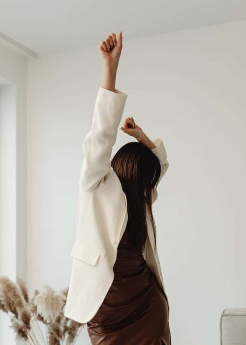 a woman stretching with one arm raised, dressed in a stylish beige blazer and brown dress, suggesting a break or moment of relaxation in a modern workspace